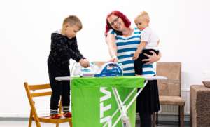 Kotipalveluyrittäjä Maarit Halonen seisoo silityslaudan takana ja pitää sylissään 2-vuotiasta Aleksi-poikaa. 4-vuotias Saku seisoo tuolilla ja silittää.